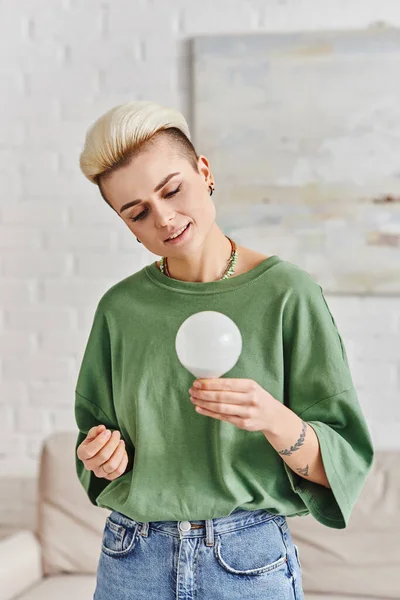 Green living, young tattooed woman with trendy hairstyle and happy face holding energy saving light bulb at home, sustainable lifestyle and environmentally conscious concept — Stock Photo