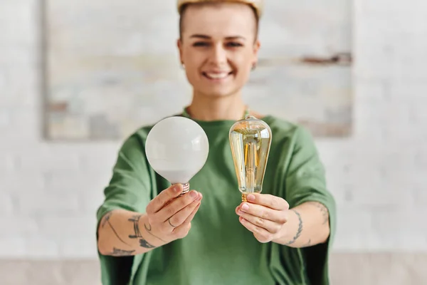 Cheerful, young and tattooed woman in casual clothes showing energy saving light bulbs and looking at camera, environmentally conscious, sustainable lifestyle and environmentally conscious concept — Stock Photo