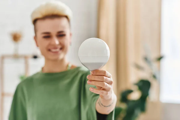 Energy saving light bulb in hand of overjoyed young woman with trendy hairstyle standing at home on blurred background, sustainable lifestyle and environmentally conscious concept — Stock Photo