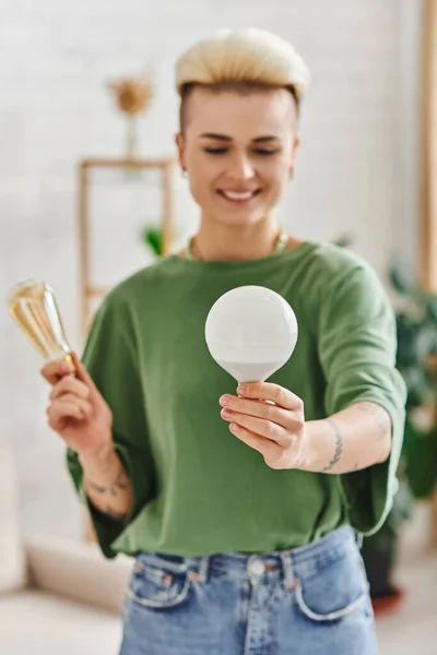 Young and tattooed woman in casual clothes holding energy saving light bulbs and smiling in living room on blurred background,  sustainable lifestyle and environmentally conscious concept — Stock Photo