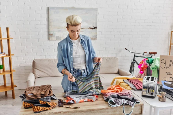 Young and tattooed woman holding knitted tank top near wardrobe items, electric toaster and cezve while preparing pre-loved items for exchange, sustainable living and mindful consumerism concept — Stock Photo