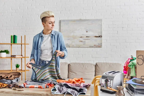 Sharing economy, tattooed and stylish woman with knitted tank-top near table with second-hand clothing and electric toaster, belongings exchange, sustainable living and mindful consumerism concept — Stock Photo