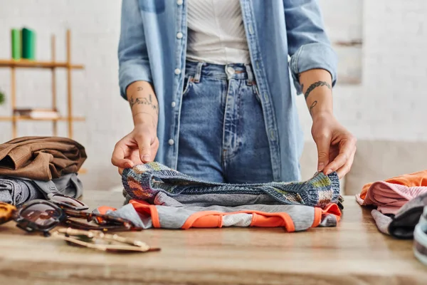 Partial view of young and tattooed woman in casual clothes sorting wardrobe items on table in living room, exchange market, eco-friendly swaps, sustainable living and mindful consumerism concept — Stock Photo