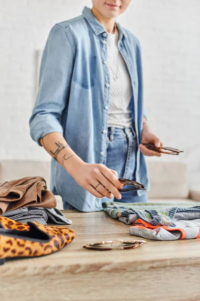 Sharing economy, swap, partial view of smiling tattooed woman in casual clothes holding sunglasses while sorting pre-loved items, sustainable living and mindful consumerism concept — Stock Photo