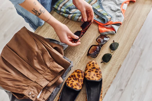 Eco-friendly swaps, top view of cropped tattooed woman near sunglasses, animal print shoes and second-hand clothing on table at home, sustainable living and mindful consumerism concept — Stock Photo