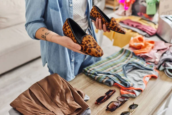 Ethical consumption, exchange, cropped view of young tattooed woman holding stylish animal-print shoes near sunglasses and garments on table, sustainable living and mindful consumerism concept — Stock Photo