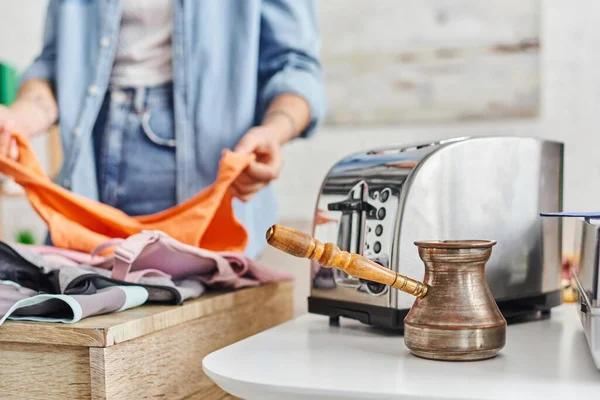 Partial view of young woman sorting clothes near electric toaster and cezve, eco-friendly swaps, exchange market, blurred background, sustainable living and promoting circular economy concept — Stock Photo