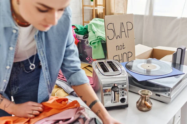 Young tattooed woman sorting second-hand clothes near electric toaster, cezve, vinyl record player and swap not shop card, blurred foreground, sustainable living and circular economy concept — Stock Photo