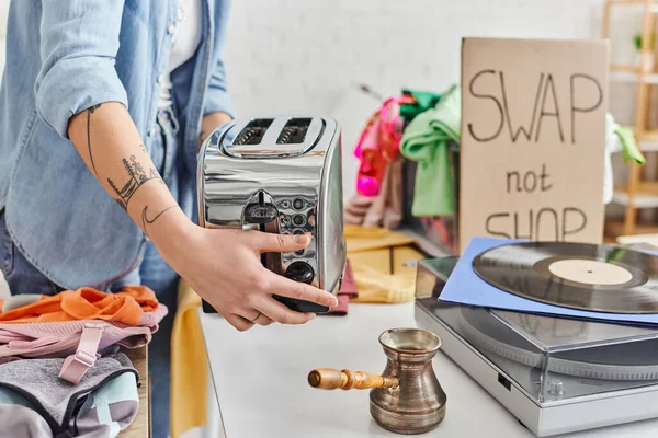 Partial view of young and tattooed woman holding electric toaster near vinyl record player, cezve and second-hand clothes during swap not shop event, sustainable living and circular economy concept — Stock Photo