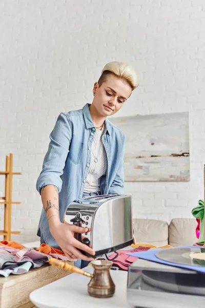Collaborative consumption, swap, smiling tattooed woman in casual clothes holding electric toaster near cezve and pre-loved clothes at home, sustainable living and promoting circular economy concept — Stock Photo