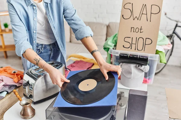 Cropped view of tattooed woman holding vinyl disc near record player, electric toaster, cezve, plastic container with clothes and swap not shop card, sustainable living and circular economy concept — Stock Photo