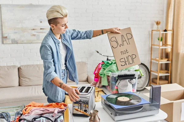 Tattooed woman with trendy hairstyle holding swap not shop card near vinyl record player, electric toaster, cezve and wardrobe clothes in living room, sustainable living and circular economy concept — Stock Photo