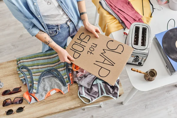 Top view of cropped tattooed woman holding swap not shop card near sunglasses, clothes, electric toaster, vinyl record player and cezve, sustainable living and circular economy concept — Stock Photo