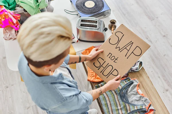 Top view of young tattooed woman holding card with swap not shop lettering near clothes, electric toaster, vinyl record player and cezve, sustainable living and circular economy concept — Stock Photo
