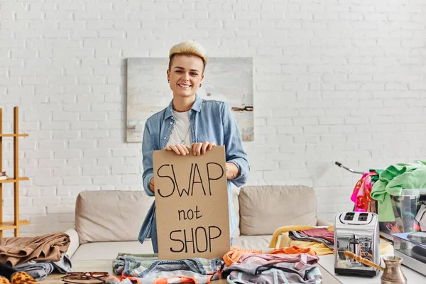 Overjoyed woman showing swap not shop card and looking at camera near second-hand clothes, electric toaster and cezve in modern living room, sustainable living and circular economy concept — Stock Photo