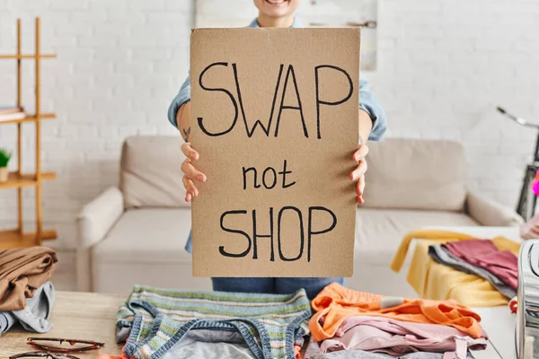Exchange market, partial view of young and cheerful woman showing card with swap not shop lettering near second-hand clothes in living room, sustainable living and circular economy concept — Stock Photo