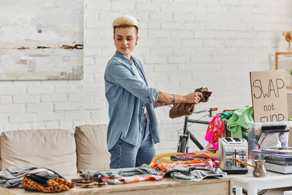 Stylish and tattooed woman holding wardrobe item near second-hand clothes, electric toaster, plastic container, cezve and swap not shop card, sustainable living and circular economy concept — Stock Photo
