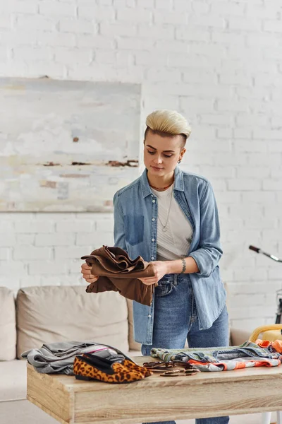 Young and tattooed woman in casual clothes, with trendy hairstyle sorting wardrobe items on table in modern living room, clothing swap, sustainable living and conscious lifestyle concept — Stock Photo