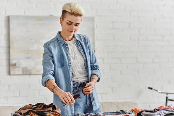 Positive tattooed woman in casual clothes, with trendy hairstyle holding sunglasses while sorting garments for exchange market, sustainable living and mindful consumerism concept — Stock Photo