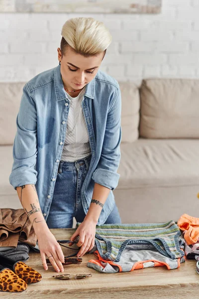 Trendy and tattooed woman placing sunglasses on table while sorting pre-loves items and clothes for swap at home, circular economy, sustainable living and mindful consumerism concept — Stock Photo