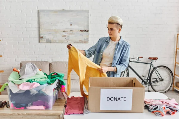Casually styled and tattooed woman holding yellow jumper while sorting clothes near plastic container and carton box with donate lettering, sustainable living and social responsibility concept — Stock Photo