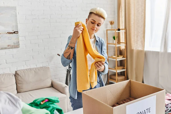 Volunteering and charity, trendy and tattooed woman standing with yellow jumper near carton box while sorting clothing for donation at home, sustainable living and social responsibility concept — Stock Photo