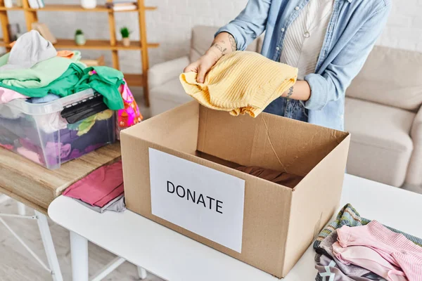 Vista parcial de una mujer joven y tatuada sosteniendo un jersey amarillo encima de una caja de cartón con letras de donaciones cerca de un contenedor de plástico con ropa, vida sostenible y concepto de responsabilidad social - foto de stock