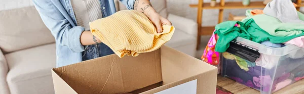 Partial view of young woman with tattoo holding yellow jumper near carton box while sorting clothes for donating for a cause, sustainable living and social responsibility concept, banner — Stock Photo
