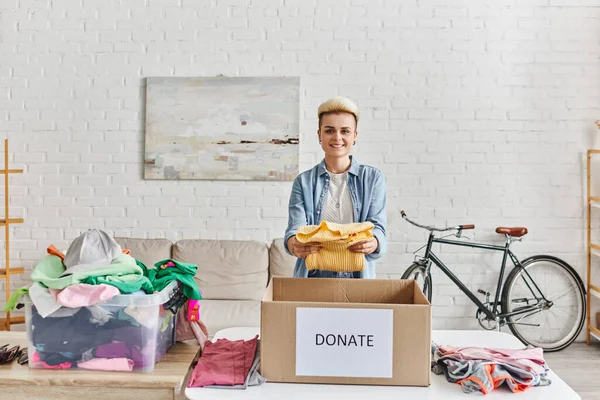 Young and positive woman with trendy hairstyle smiling at camera and holding yellow jumper near donation box and plastic container with clothes, sustainable living and social responsibility concept — Stock Photo