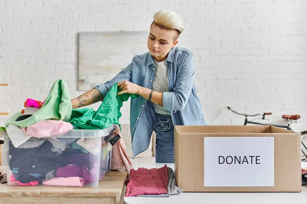 Promoting social good, young tattooed woman sorting clothing in plastic container near carton box with donate lettering in modern living room, sustainable living and social responsibility concept — Stock Photo