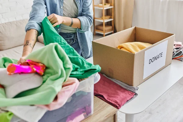 Cropped view of tattooed woman in casual clothes sorting pre-loved and second-hand items near carton box with donate lettering at home, sustainable living and social responsibility concept — Stock Photo