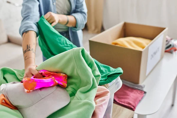 Social good initiatives, partial view of young and tattooed woman sorting casual clothes near carton box for donating for a cause, sustainable living and social responsibility concept — Stock Photo