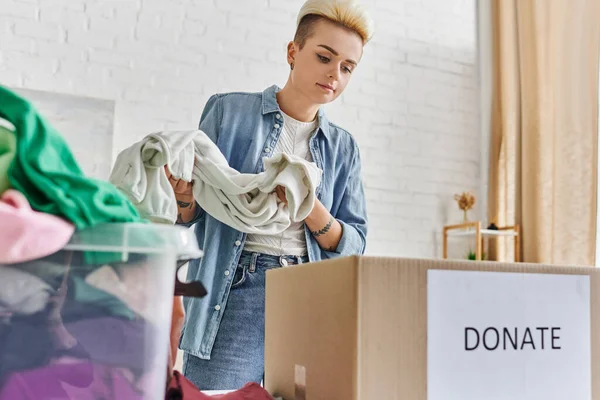 Donating for a cause, casually styled and tattooed woman holding clothes near plastic container and carton box with donate lettering, sustainable living and social responsibility concept — Stock Photo