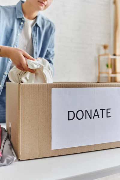 Partial view of young woman in casual clothes packing garments in carton box with donate lettering, promoting social good, sustainable living and social responsibility concept, blurred background — Stock Photo