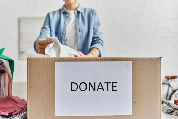 Social good initiatives, partial view of young woman packing clothing in carton box with donate lettering at home, blurred background, sustainable living and social responsibility concept — Stock Photo