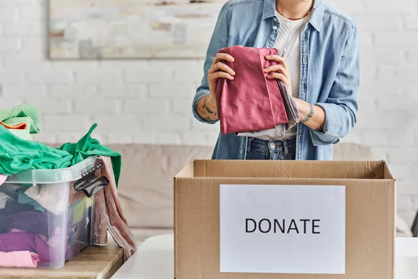 Charity and volunteering, cropped view of young and tattooed woman holding wardrobe garments near carton box with donate lettering, sustainable living and social responsibility concept — Stock Photo