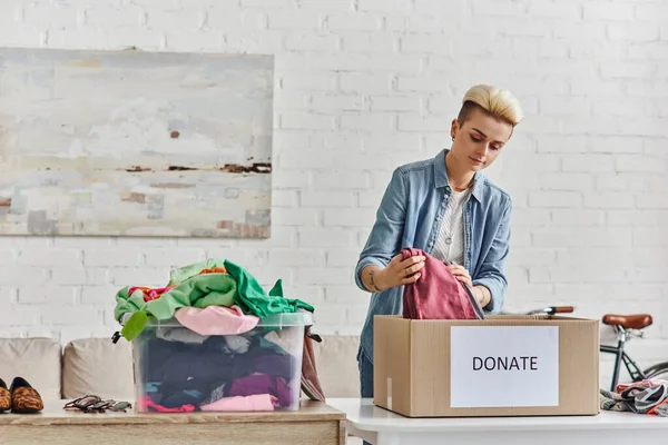 Spenden für einen guten Zweck, stylische tätowierte Frau verpackt Kleidungsstücke in Spendenbox in der Nähe von Plastikbehältern mit Second-Hand-Artikeln im Wohnzimmer, nachhaltiges Leben und soziales Verantwortungskonzept — Stockfoto