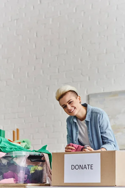 Young and joyful woman with trendy hairstyle holding pre-loved clothes and looking at camera near plastic container and donation box at home, sustainable living and social responsibility concept — Stock Photo