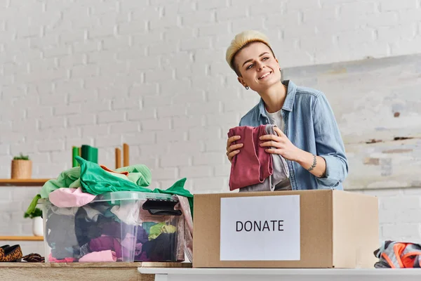 Cheerful tattooed woman looking away while standing with clothes near donation box and plastic container with wardrobe items at home, sustainable living and social responsibility concept — Stock Photo