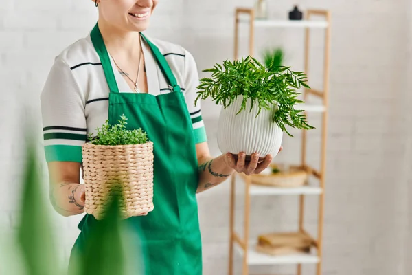 Indoor gardening, partial view of happy tattooed woman in green apron holding flowerpots with houseplants in modern living room, blurred foreground, sustainable home decor and green living concept — Stock Photo