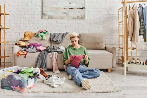 Young tattooed woman in casual clothes sorting thrift store finds while sitting on floor near couch in modern living room, sustainable living and mindful consumerism concept — Stock Photo