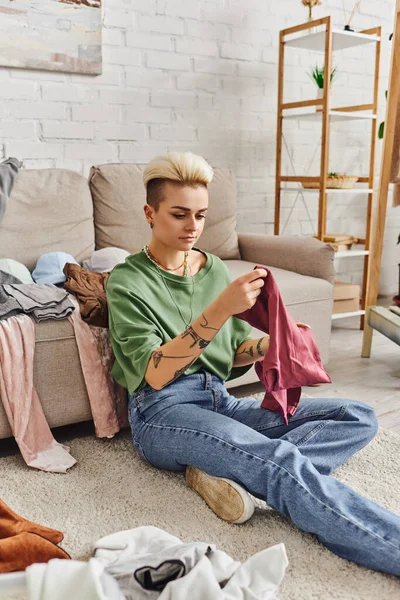 Casually styled and tattooed woman with trendy hairstyle sitting on floor and sorting second-hand items near couch in modern living room, sustainable living and mindful consumerism concept — Stock Photo
