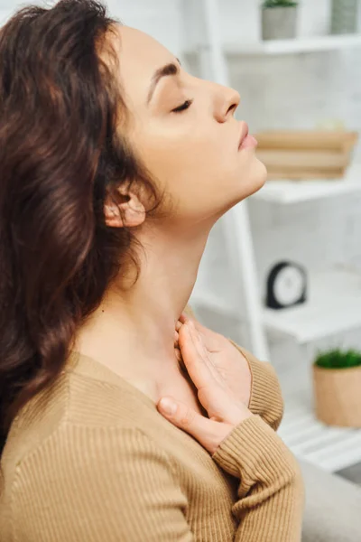 Vue latérale de la jeune femme brune en pull occasionnel touchant le cou lors d'un massage à domicile du système lymphatique dans une maison floue, un rituel d'autosoin et un concept holistique de pratiques de bien-être — Photo de stock