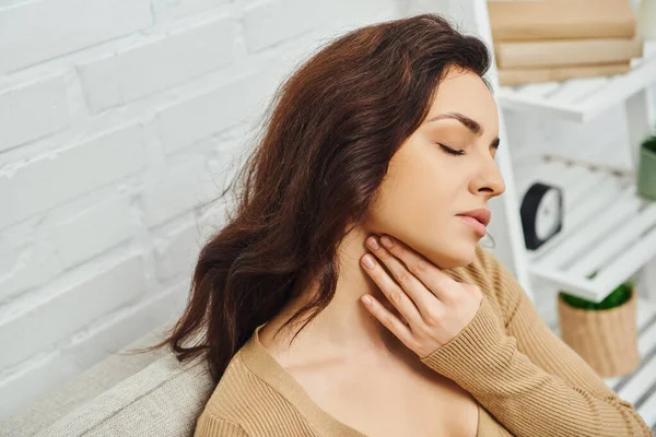 High angle view of young brunette woman in casual jumper massaging lymphatic nodes on neck while sitting on couch at home, self-care ritual and holistic wellness practices concept, tension relief — Stock Photo