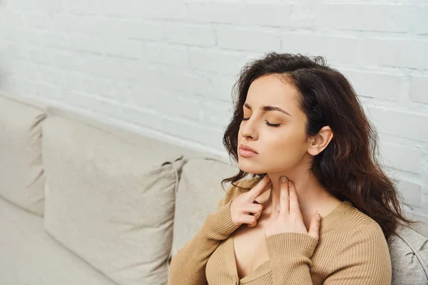 Young brunette woman with closed eyes in casual jumper massaging lymphatic nodes on neck during self-massage while sitting on couch at home, self-care ritual and holistic wellness practices concept — Stock Photo