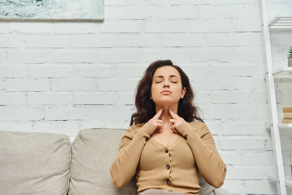Young brunette woman with closed eyes in brown jumper touching neck during lymphatic nodes and circulation massage on couch at home, self-care ritual and holistic wellness practices concept — Stock Photo