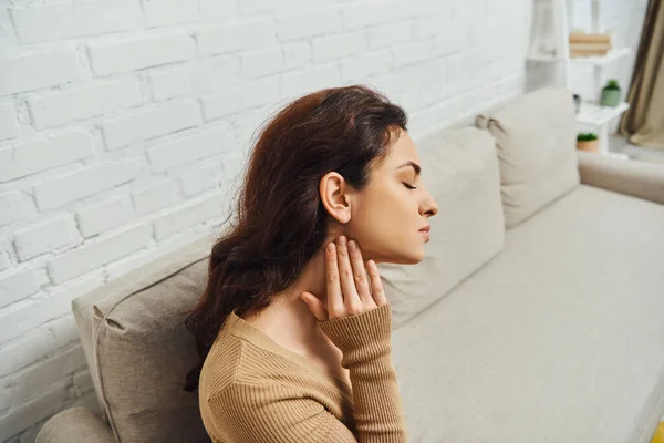 Side view of young brunette woman in brown jumper massaging neck during massage for lymphatic circulation and sitting on couch at home, self-care ritual and holistic wellness practices concept — Stock Photo