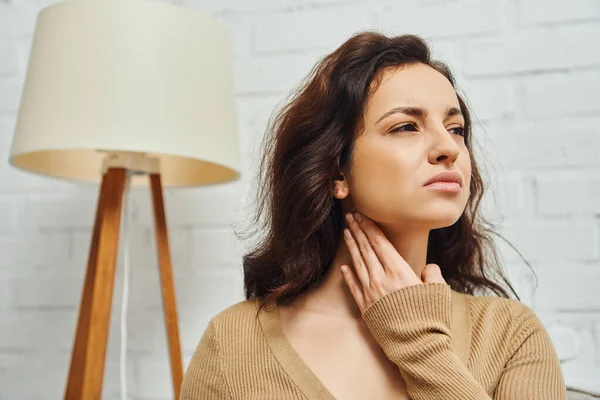 Displeased young brunette woman in brown jumper touching neck during lymphatic nodes self-massage at home, self-care ritual and holistic wellness practices concept, tension relief — Stock Photo