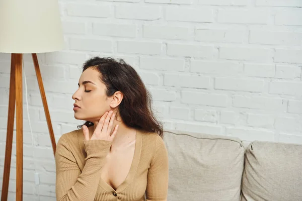 Vue latérale de la jeune femme brune en pull brun massant le cou pendant le massage lymphatique à domicile et assise sur le canapé, concept de guérison holistique et rituel d'autosoin, soulagement de la tension — Photo de stock