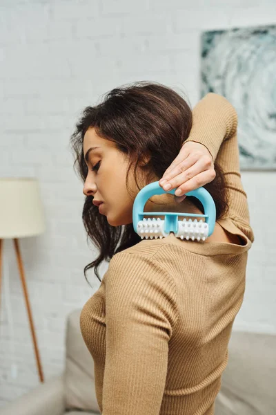 Side view of young brunette woman in brown jumper using handled massager while doing self-massage on shoulder at home, enhancing self-awareness and body relaxation concept, balancing energy — Stock Photo
